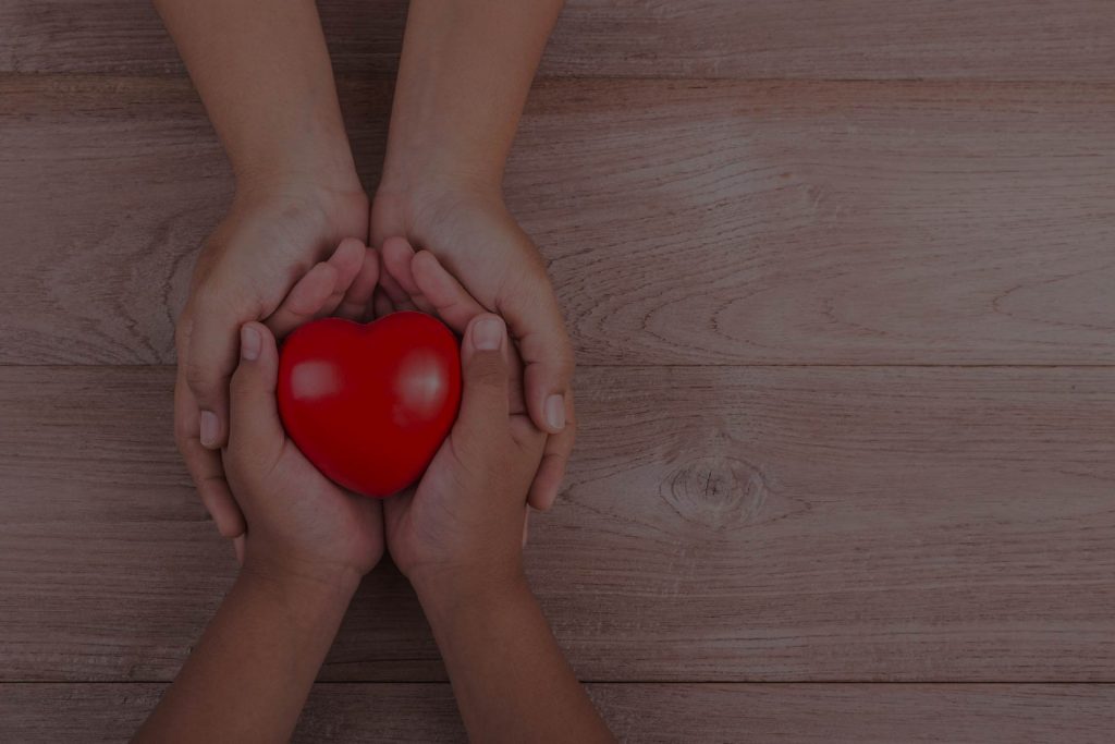 Två par händer som tillsammans håller ett rött hjärta – symbol för omtanke, stöd och välgörenhet.